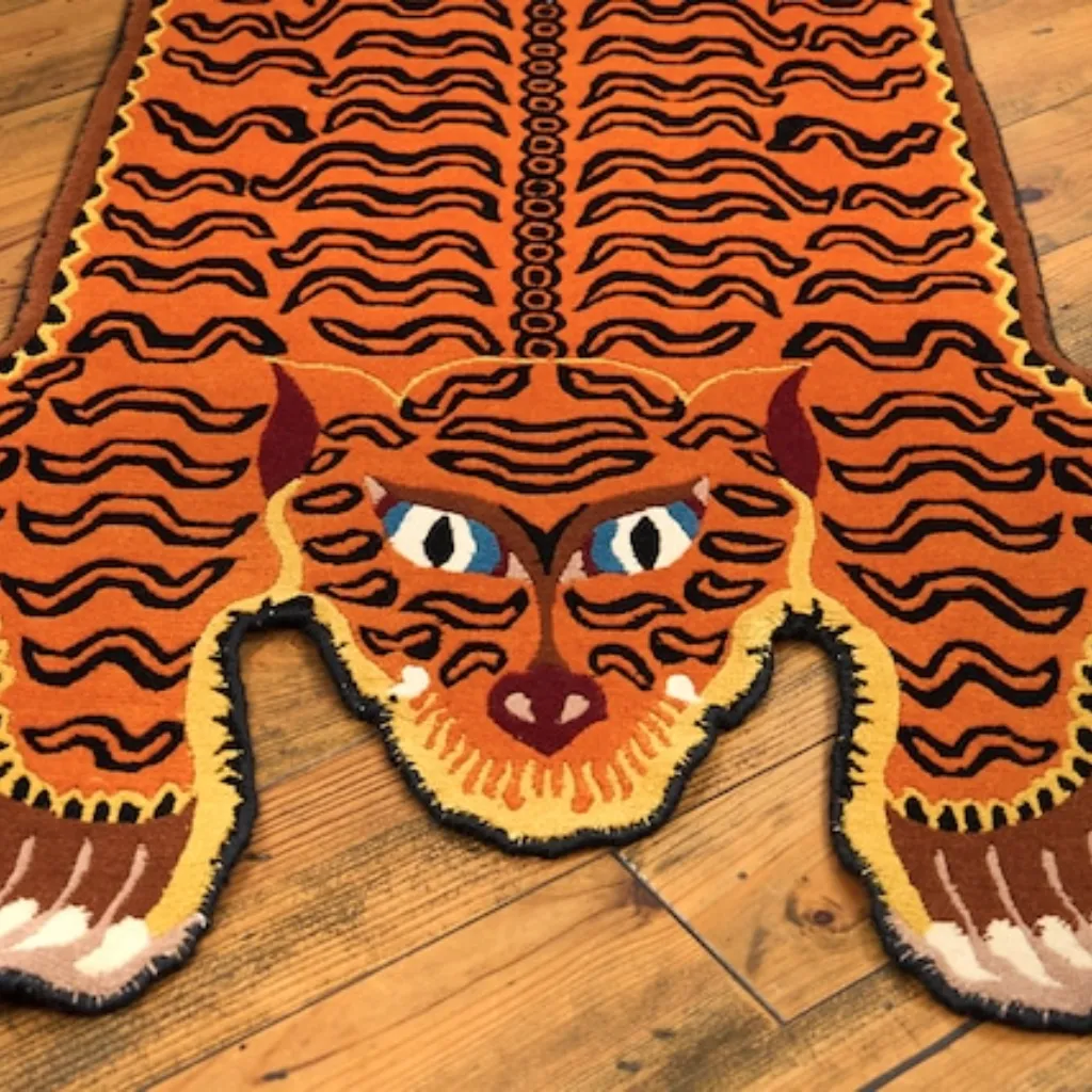 amber raja tibetan tiger rug head detail Close-up top down view of the Amber Raja Tibetan Rug, focusing on the stylized tiger face with vibrant blue eyes, orange fur, and black wavy stripes.