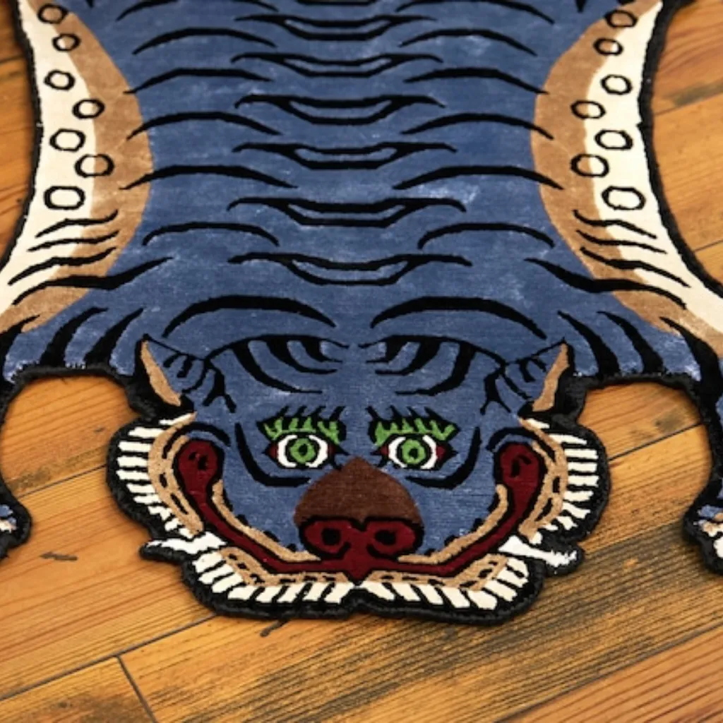 ashen claw tibetan blue grey tiger rug head detail Close-up top down view of the Ashen Claw Tibetan Rug, focusing on the stylized blue-grey tiger face with striking green eyes, brown nose, and black wavy stripes.