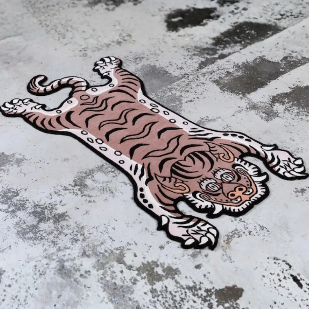 blush prowl hand tufted wool tiger rug design Close-up top down view of the Blush Prowl Tibetan Rug, focusing on the stylized tiger face with striking teal eyes, sharp teeth, and black wavy stripes on a soft blush pink background.