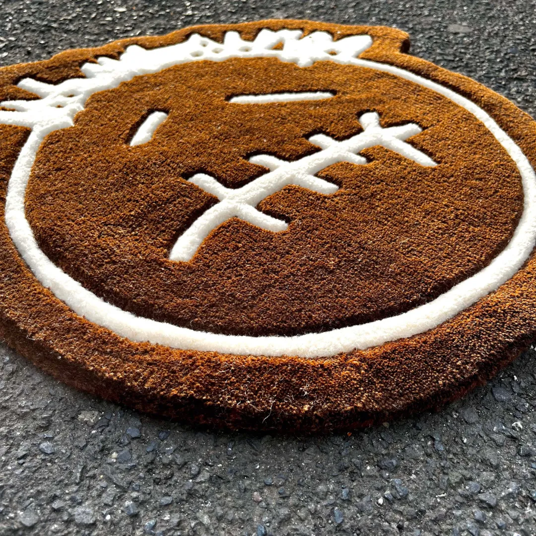 Close-up angled view of the Bruma Mark rug placed on a dark asphalt surface, highlighting the dense, plush brown hand-tufted wool texture and the sharply carved white lines forming the stitched mouth detail.