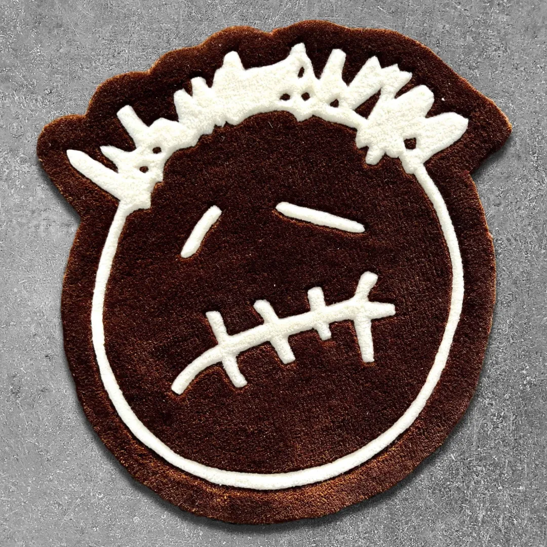 Top-down view of the shaped Bruma Mark rug on a textured grey surface, featuring a solid brown base with thick white embroidered-style lines forming a stylized face with closed eyes, a stitched mouth, and spiky hair.