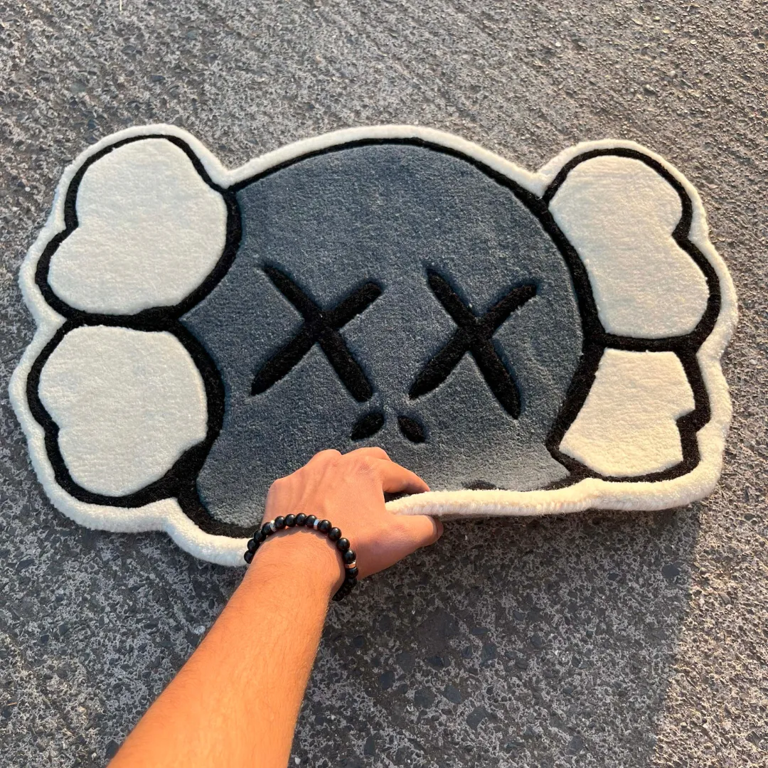 Angled view showing a hand lifting the bottom edge of the Gravine Emblem rug on an asphalt street, emphasizing the thick, flexible wool pile and the bold black outlines of the skull design.