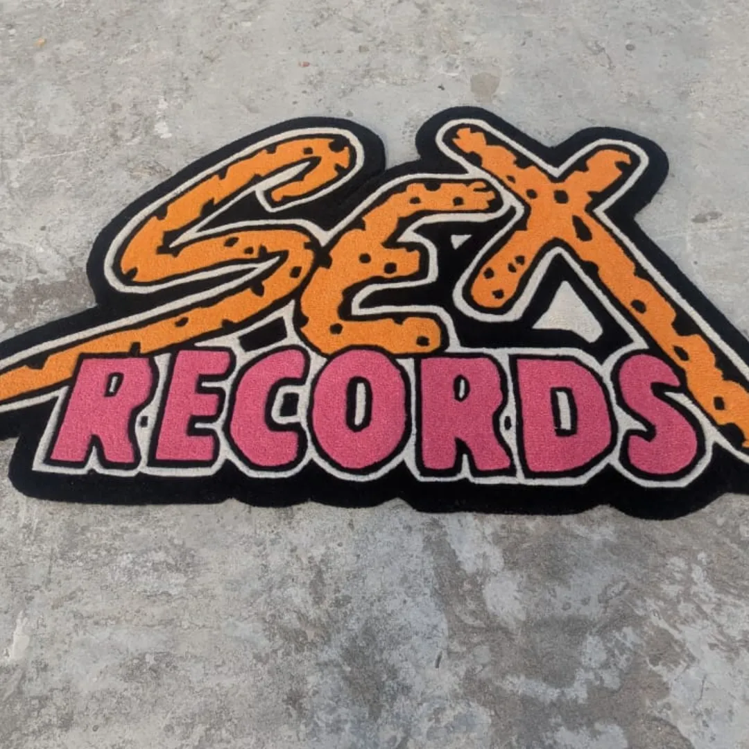 Slightly angled view of the Neon Graffiti rug on a concrete background, showcasing its unique text-based silhouette and the striking contrast of the thick black outline against the bright orange and pink letters.