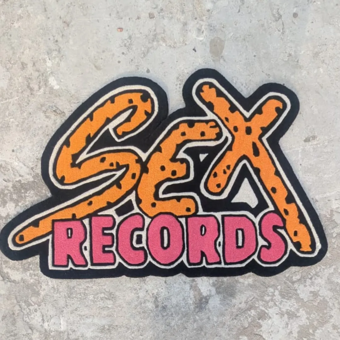 Top-down view of the shaped Neon Graffiti rug placed on a concrete surface, featuring bold pop-art typography that reads "SEX RECORDS" with orange and black cheetah print top letters and vibrant pink bottom letters.