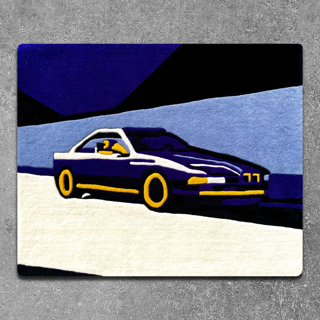 Top-down view of the rectangular Noctra Veil rug placed on a concrete road, featuring a bold, stylized profile of a retro blue and white sports car against a color-blocked dark and light blue background.