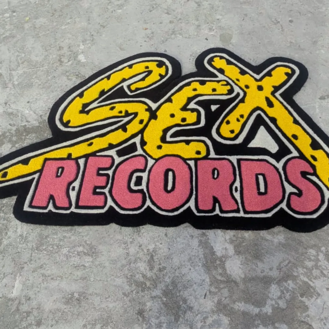 Slightly angled view of the Vault Graffiti Drop rug on a concrete background, showcasing its unique text-based silhouette and the striking contrast of the thick black outline against the bright yellow and pink letters.