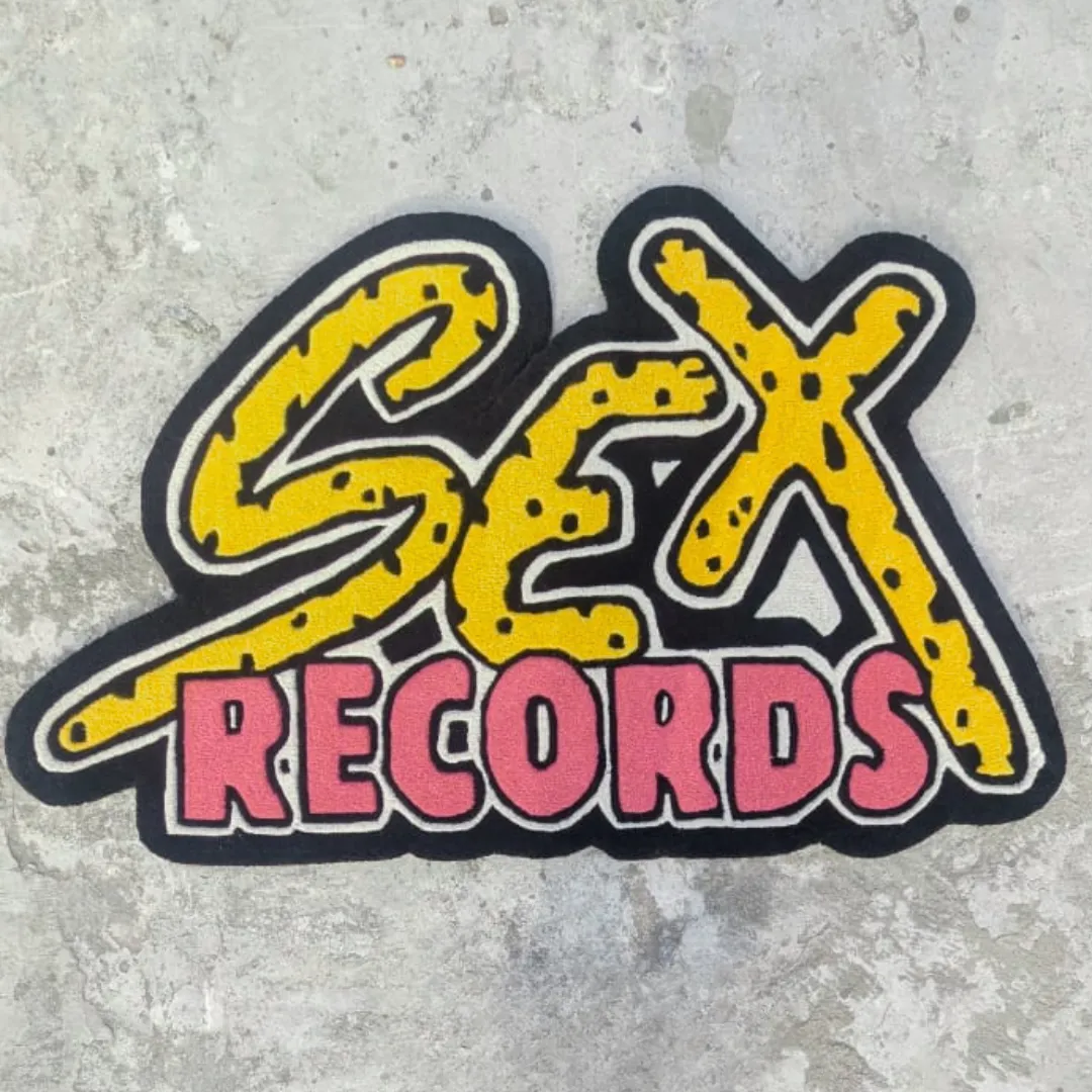 Top-down view of the shaped Vault Graffiti Drop rug placed on a concrete surface, featuring bold pop-art typography that reads "SEX RECORDS" with yellow and black cheetah print top letters and vibrant pink bottom letters.