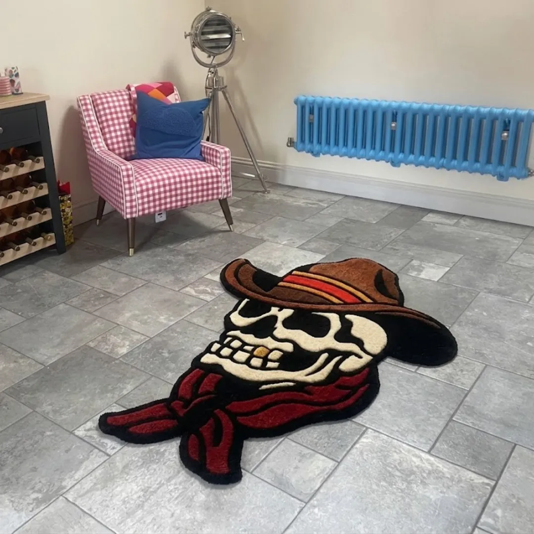 An angled view of the hand-tufted Skull Cowboy rug placed on a grey tiled floor. The classic American traditional tattoo-style design features a skull with a gold tooth, wearing a brown cowboy hat and a vibrant red bandana, styled in a room next to a pink checkered armchair and a bright blue radiator.