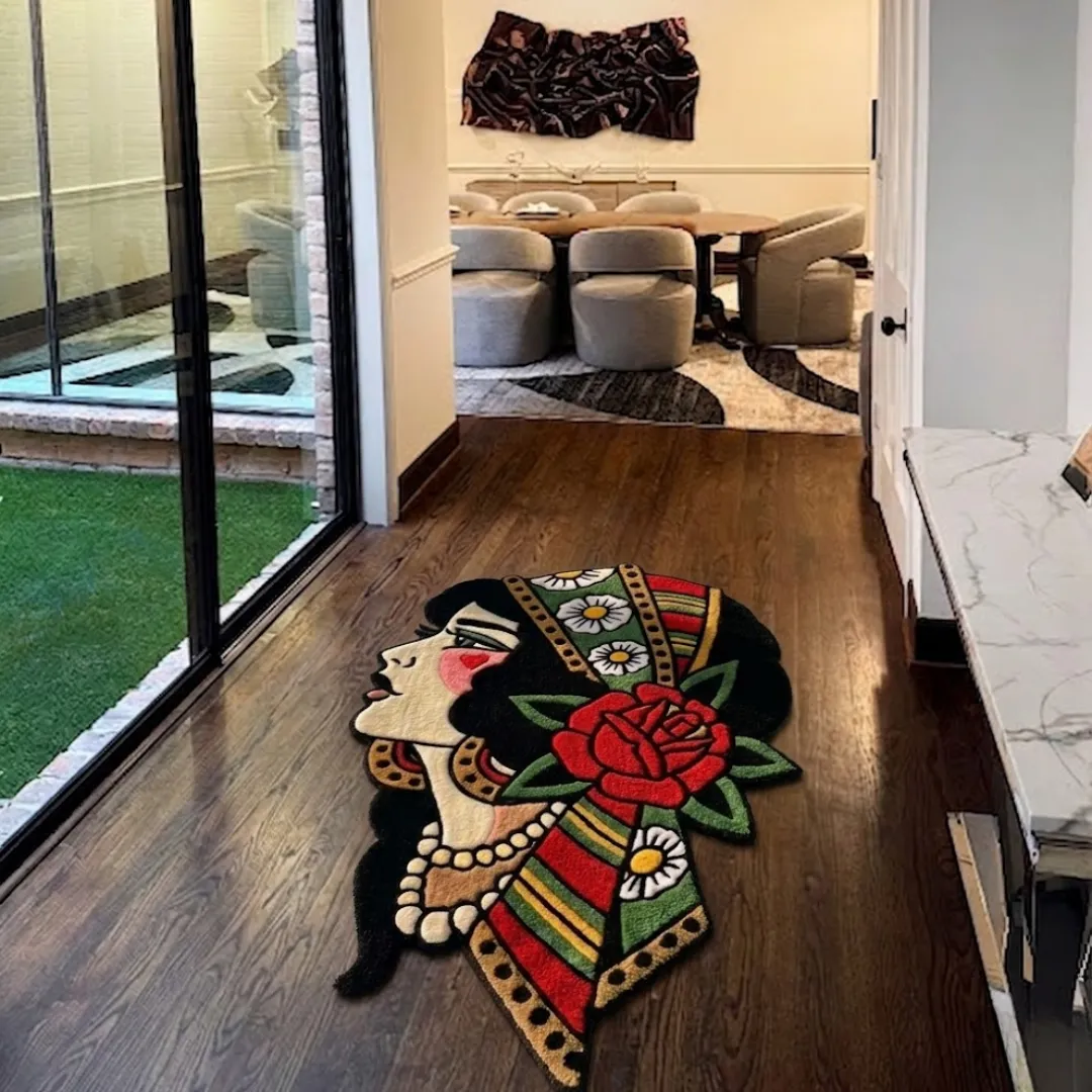 An angled view of the Lady Rose Flash Art rug resting on a dark hardwood floor in a modern home hallway. The hand-tufted custom rug features a classic American traditional tattoo design of a woman's profile with dark hair, a pearl necklace, a large red rose, and a colorful green, red, and yellow headscarf detailed with white daisies. The rug is positioned near sliding glass doors, leading into a contemporary dining room.