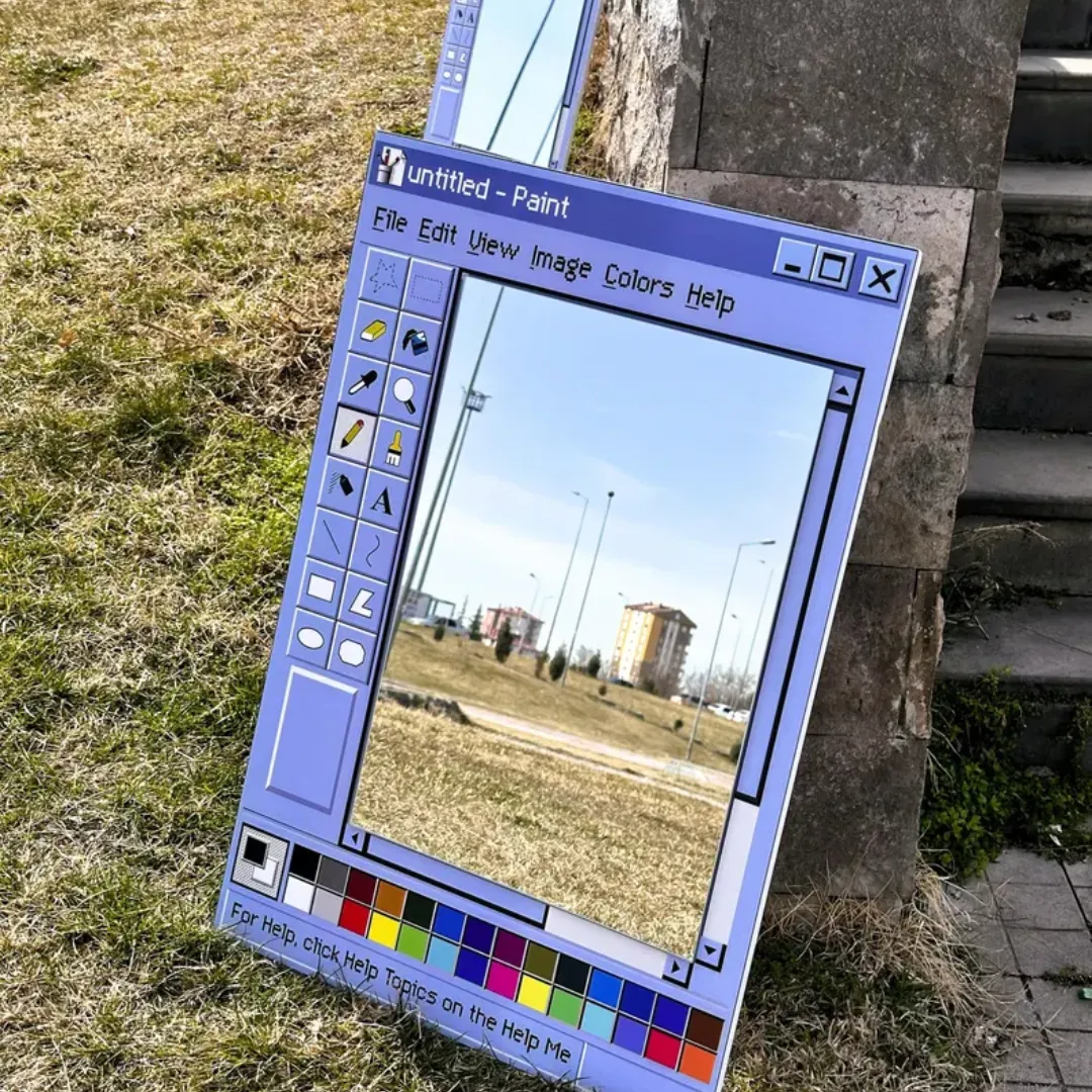 retro window paint app mirror garden display An outdoor display of a retro window mirror styled as a computer paint application, resting on a grassy lawn beside a stone structure.