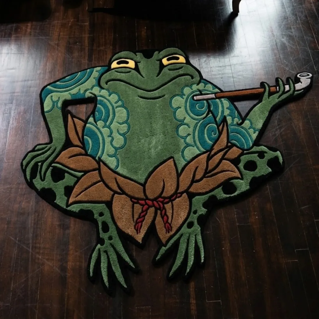kaeru musashi japanese tattoo rug dark wood floor An overhead view of the Kaeru Musashi rug resting on a dark hardwood floor. The hand-tufted custom rug features a traditional Japanese tattoo design of a relaxed green frog with yellow eyes and intricate blue wave tattoos on its arms. The frog is holding a traditional smoking pipe and wearing a brown leaf skirt tied with a red braided cord.