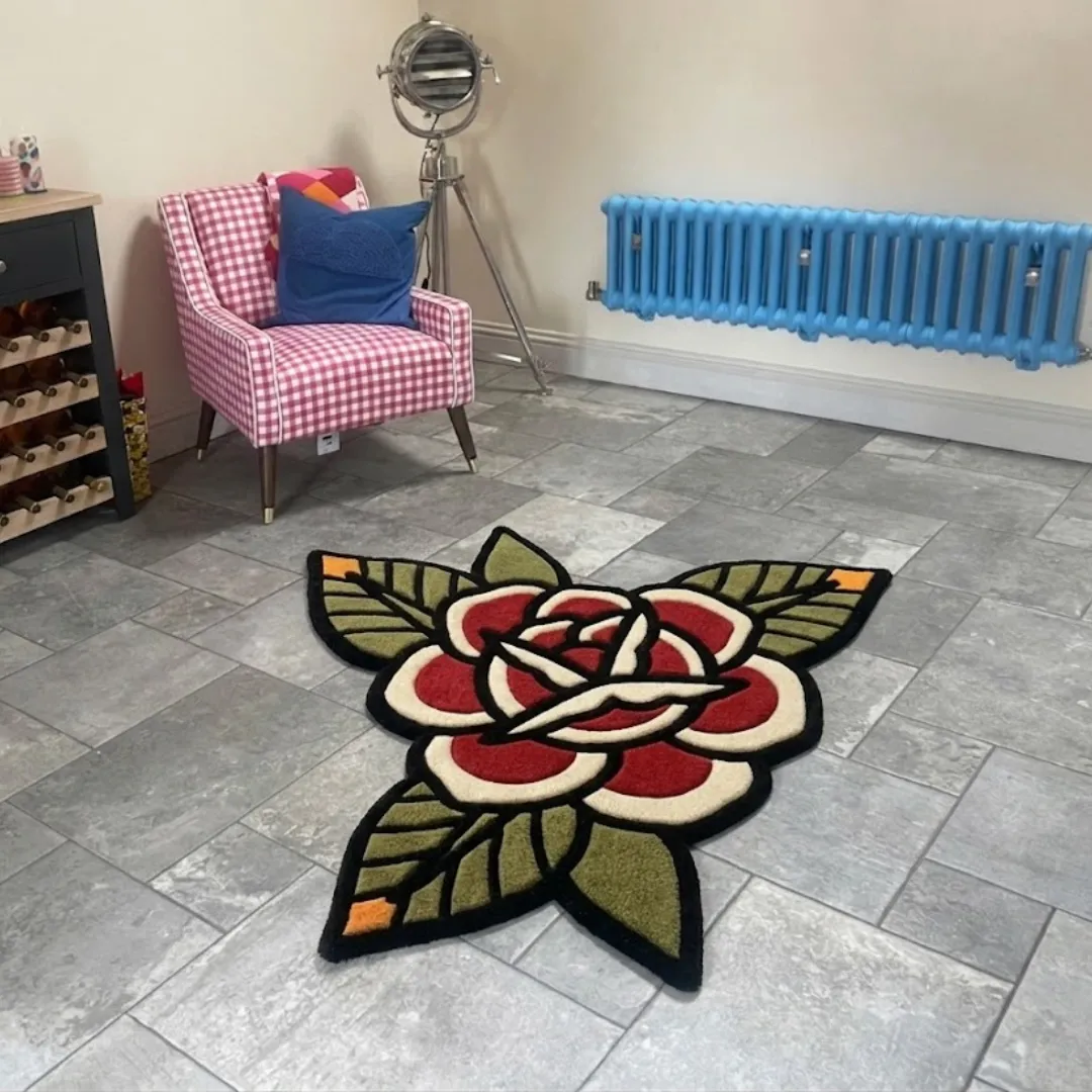 red rose flower traditional rug room setting An angled view of the Red Rose Flower Rug placed on a grey tiled floor. The hand-tufted design features a classic American traditional tattoo style with a bold red blossom, cream-colored inner petals, and green leaves outlined in black, styled in a room next to a pink checkered armchair and a bright blue radiator.