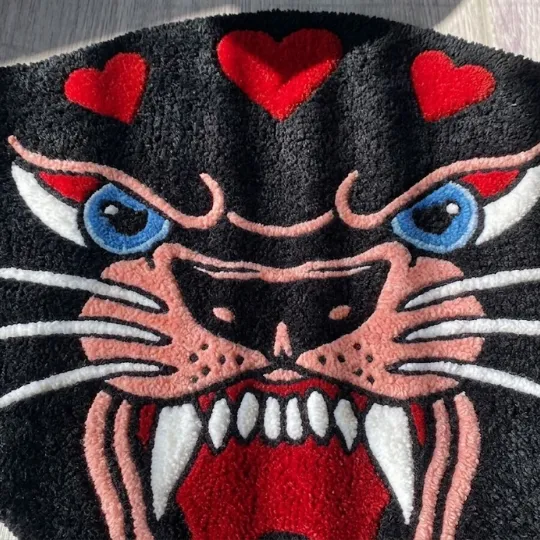 panther head rug close up A full overhead shot of the Panther Head rug resting in a patch of sunlight on a light-colored floor. The design showcases a fierce black panther silhouette with pink facial details, sharp teeth, and red hearts across the top.