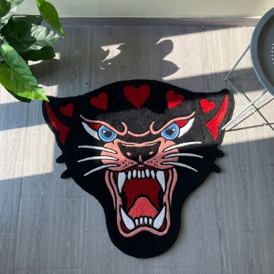 panther head rug light wood floor A full overhead shot of the Panther Head rug resting in a patch of sunlight on a light-colored floor. The design showcases a fierce black panther silhouette with pink facial details, sharp teeth, and red hearts across the top.