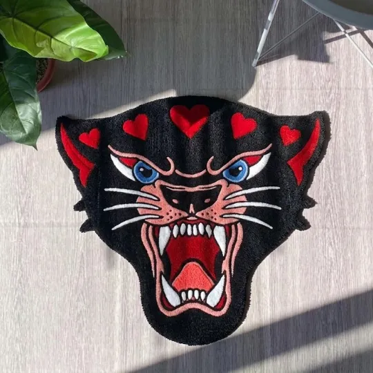 panther head rug top down view Top-down view of the tufted Panther Head rug placed on a light wood floor next to a green houseplant. The rug features a traditional tattoo-style black panther face with an open roaring mouth, blue eyes, and red heart accents on the forehead.