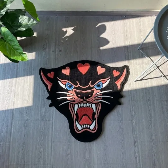 pink panther head rug full view Full view of a tufted black panther head rug with pink hearts, blue eyes, and a roaring mouth on a sunlit wooden floor.