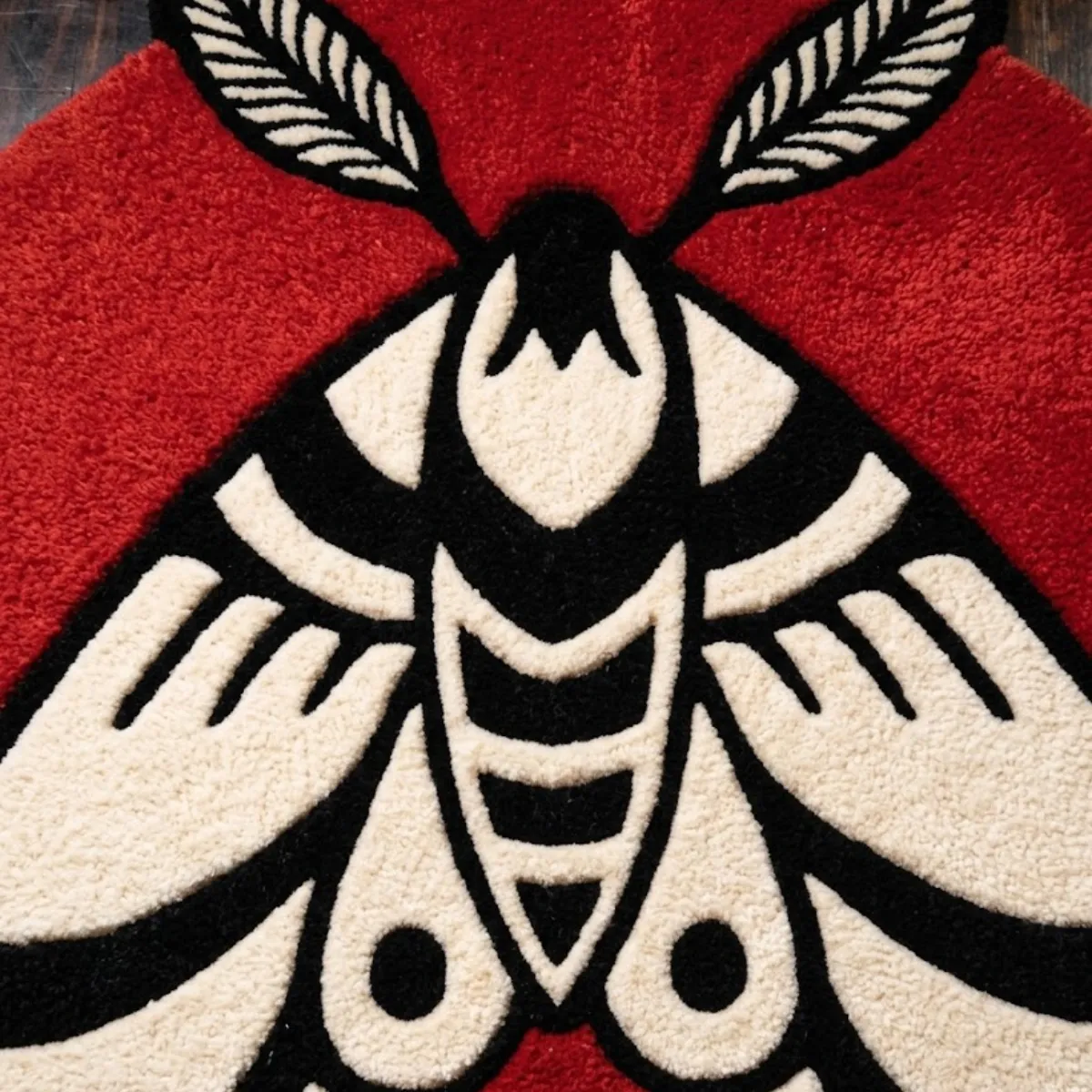 A close-up, top-down view of the Moth Round Rug resting on a dark wood floor. The detailed shot highlights the moth's geometric body and feathered antennae, emphasizing the crisp black outlines and the thick, tufted New Zealand wool and viscose yarn, crafted with internal stuffing for a luxurious, natural softness.