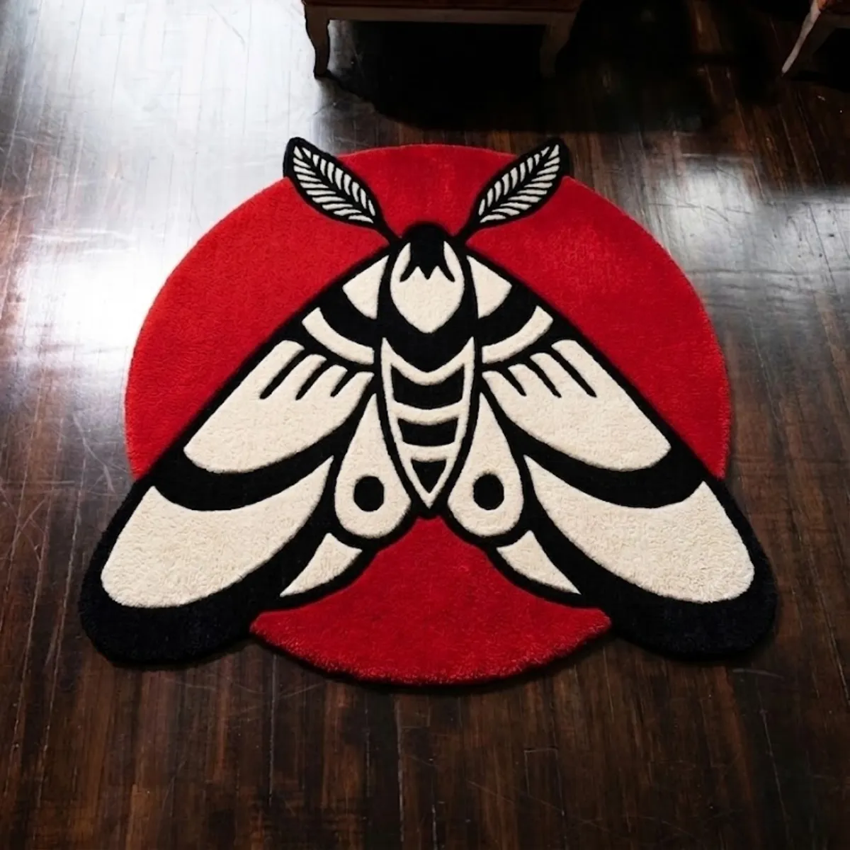 An overhead view of the Moth Round Rug resting on a dark hardwood floor. The hand-tufted rug features a bold traditional tattoo-style design of a black and cream moth with flat, unshaded color blocking, set against a vibrant solid red circular background.