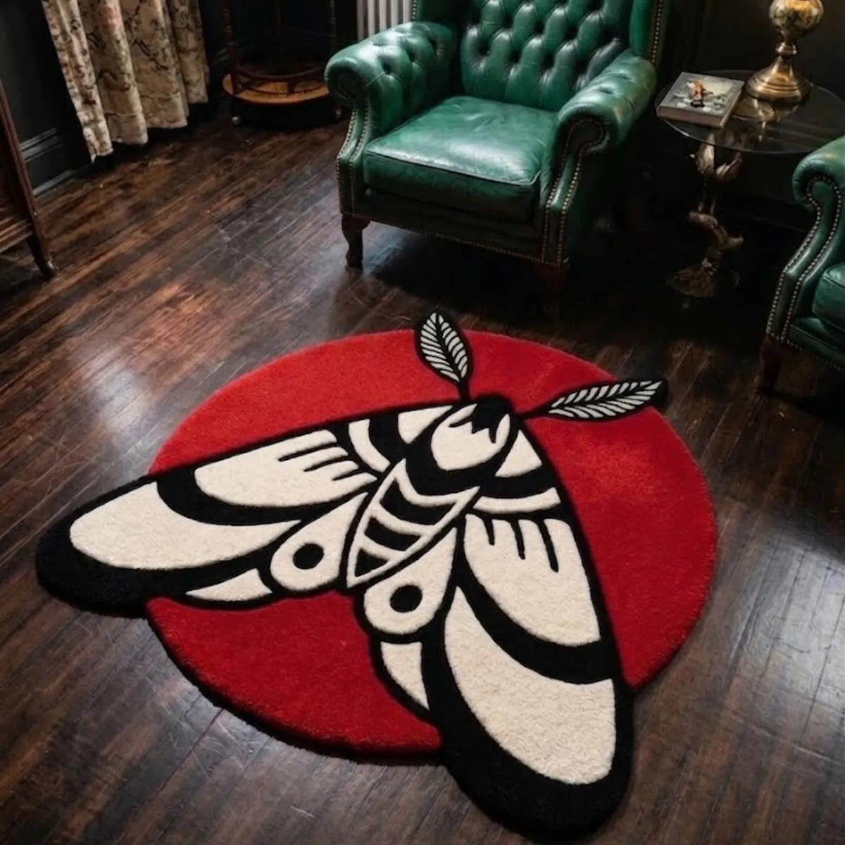 An angled view of the custom-shaped Moth Round Rug placed on a dark wood floor in a moody, classic room setting. The bold black, cream, and red moth design partially extends past its circular base, styled beautifully next to vintage tufted green leather armchairs.