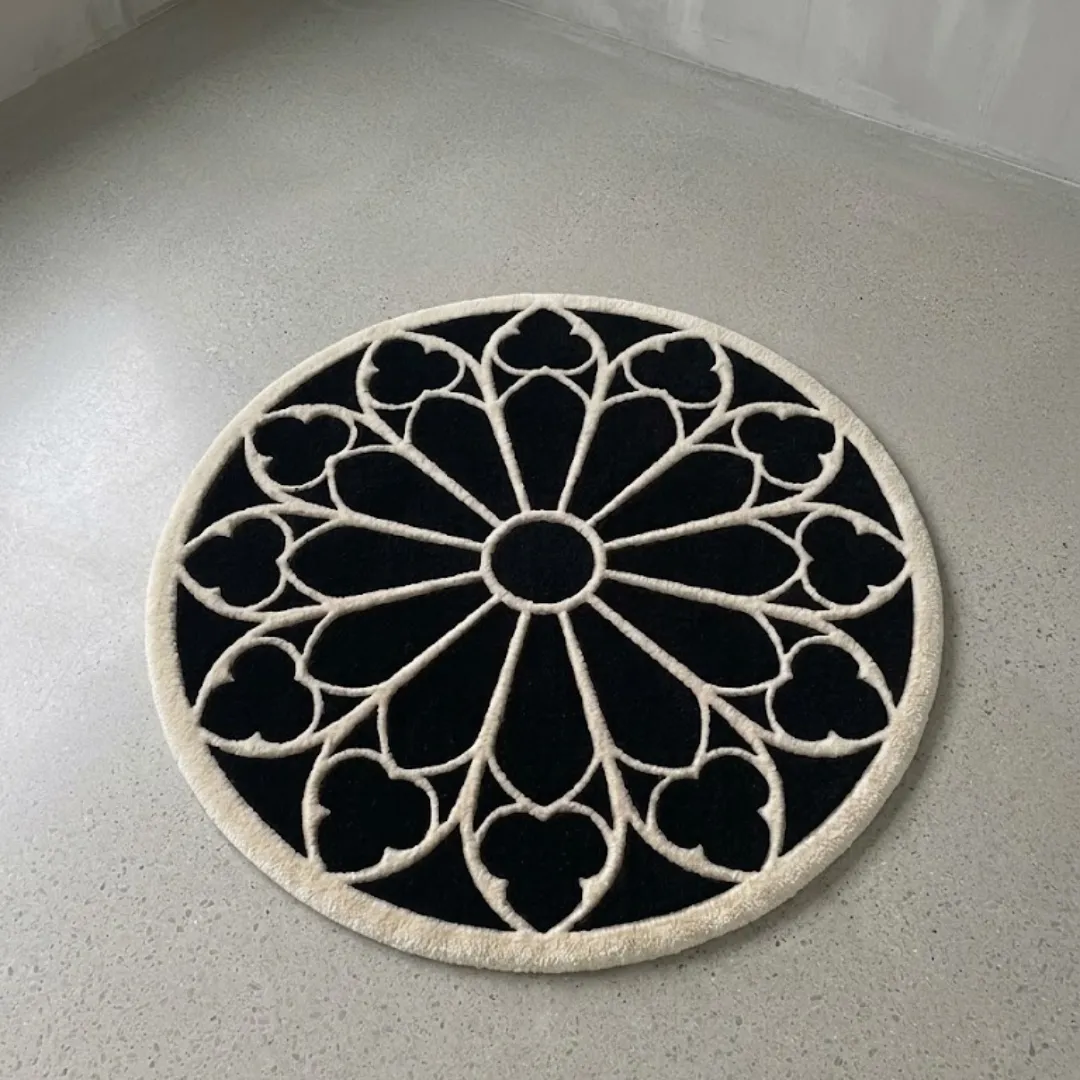 eclipse halo rug overhead view A full top-down view of the round Eclipse Halo Rug resting on a light grey concrete floor. The hand-tufted rug features a striking black background with a contrasting white geometric gothic rose window pattern and a thick white outer border.