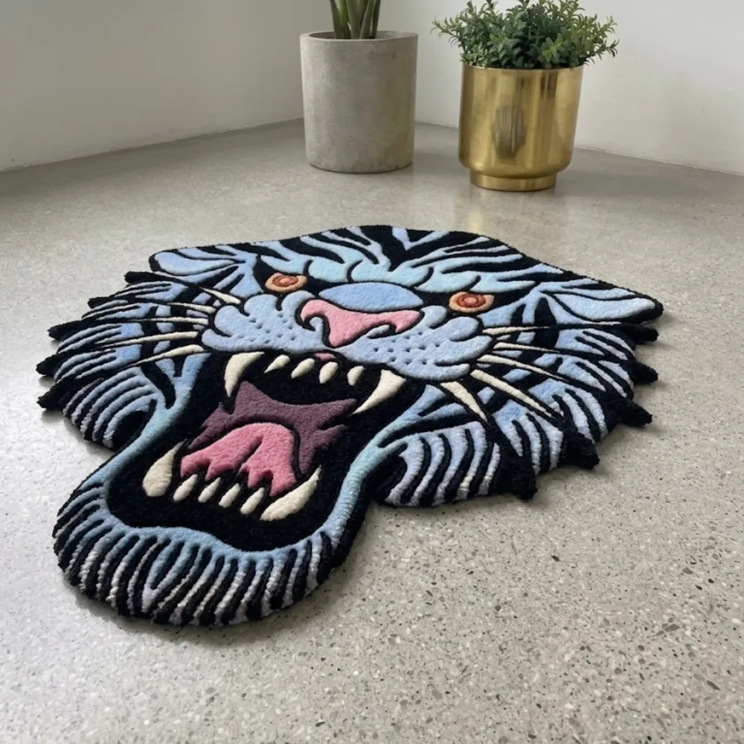 rebellion blue rug angled view An angled shot of the Rebellion Blue Rug placed on a smooth concrete floor near a gold planter. The image highlights the custom cut-out shape of the blue and black American traditional tiger head design and its thick, plush tufted texture.