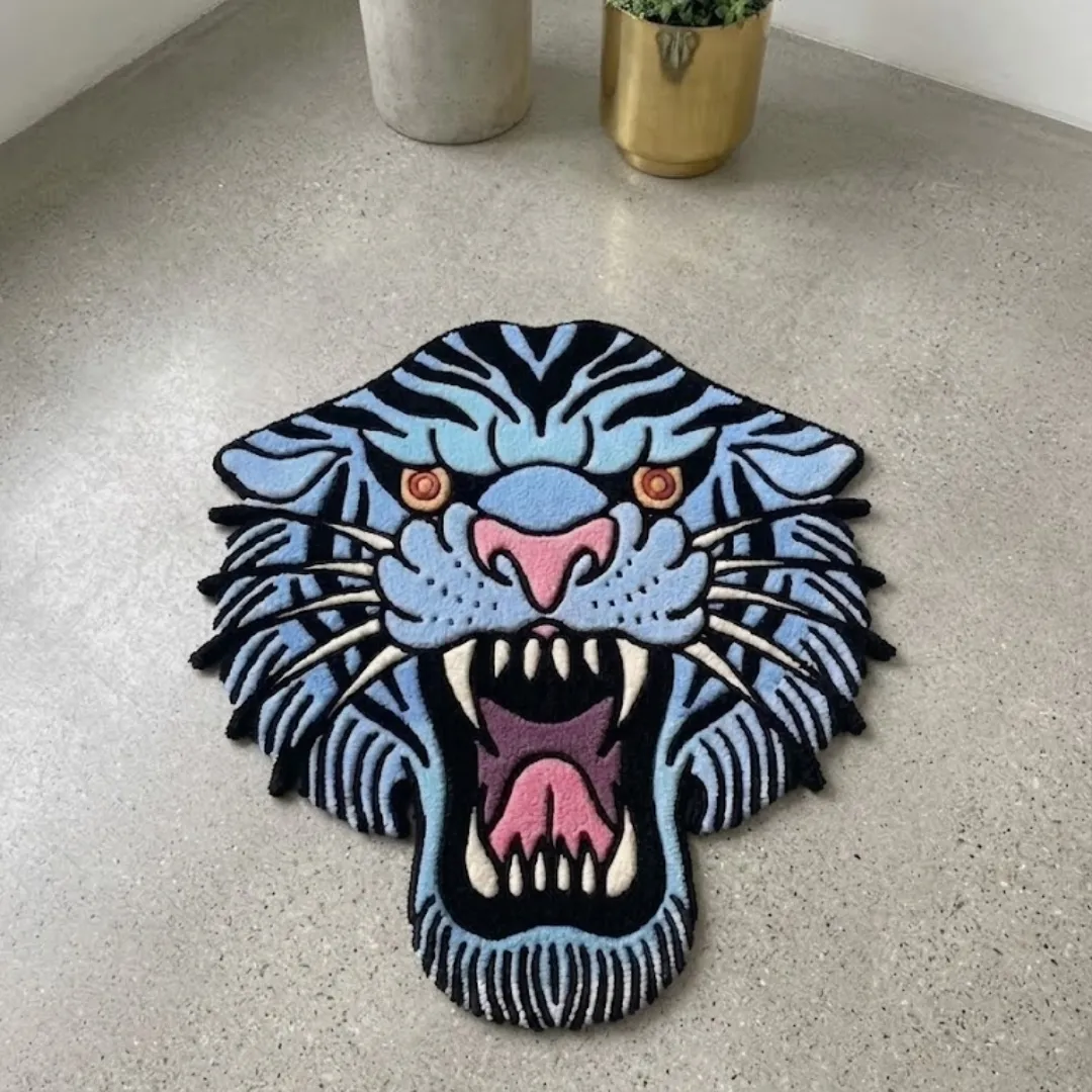 rebellion blue rug top down view A top-down view of the Rebellion Blue Rug resting on a light speckled concrete floor. The hand-tufted rug features an American traditional tattoo style tiger head in light blue and black stripes, with a fierce open mouth, pink nose, and sharp white teeth.