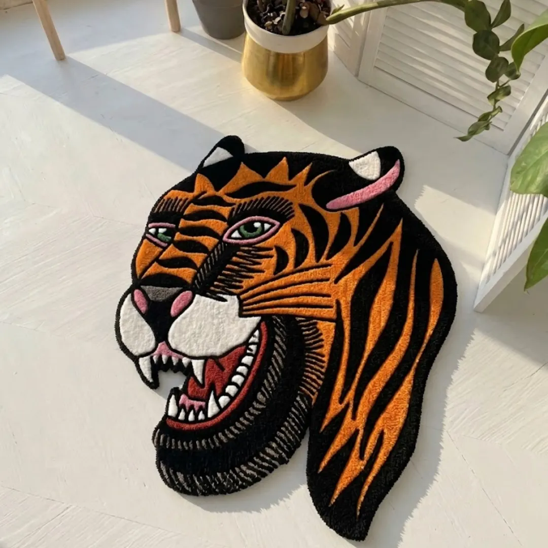 bert grimm tiger traditional rug white floor An angled view of the Bert Grimm Tiger Traditional Rug resting on a white wooden floor bathed in natural sunlight. The hand-tufted rug showcases a classic American traditional tattoo-style tiger head featuring bold red and black stripes, a crisp white muzzle, striking green eyes, and red detailing on the nose and inner ears, positioned near a gold planter and a white slatted room divider.