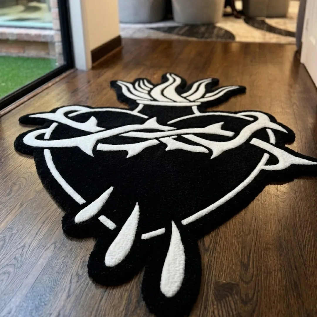 thorned heart rug close up A close-up, angled view of the hand-tufted Thorned Heart Rug resting on a dark hardwood floor. The black and white rug features a bold sacred heart design, showcasing thick white tufted yarn forming flames, a wrapping crown of thorns, and falling drops against a solid black background.