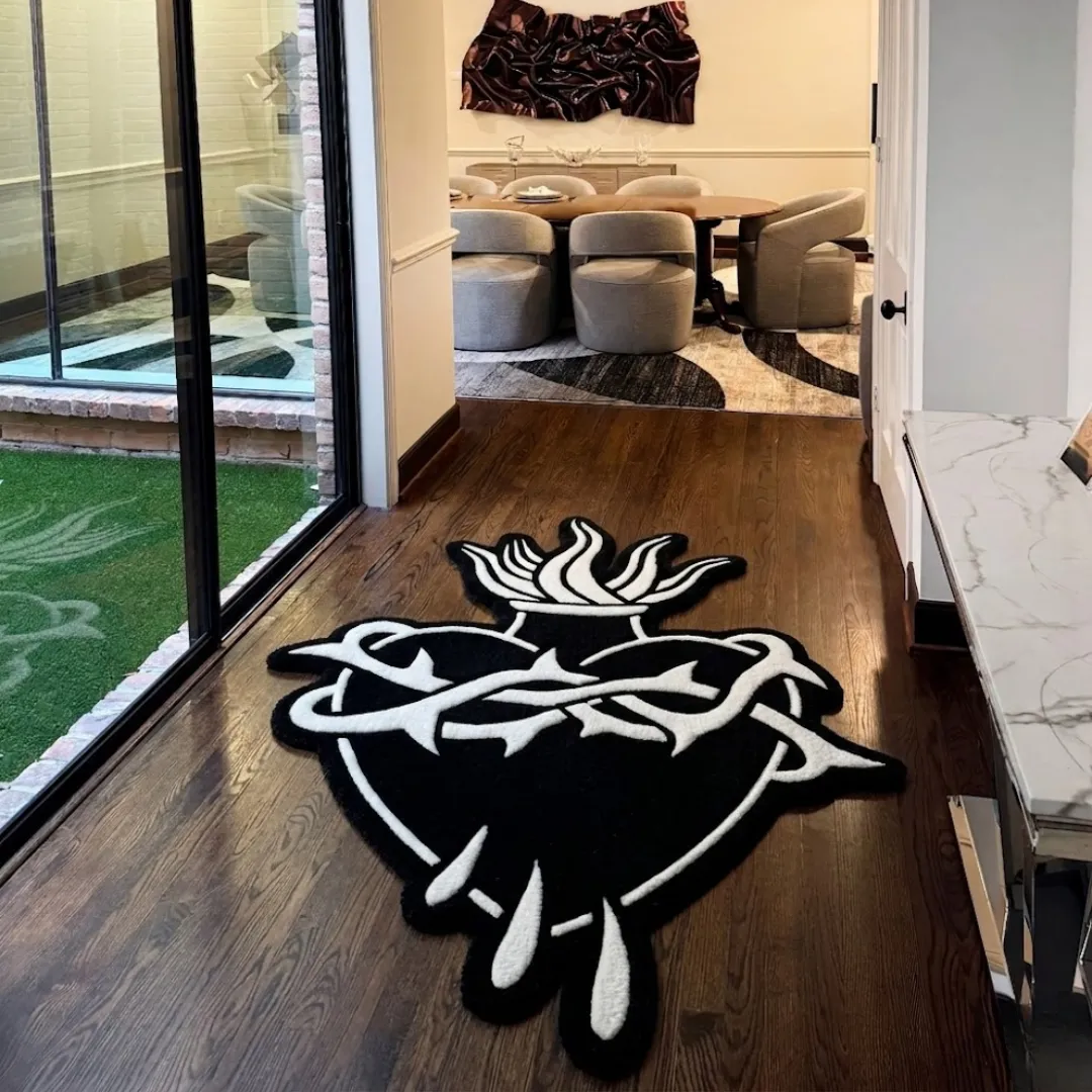 thorned heart rug hallway setting The black and white Thorned Heart Rug styled in a modern home hallway with dark wood floors. The custom cut-out tufted rug, featuring a striking sacred heart, thorns, and flames, is positioned near large glass sliding doors, leading the eye toward a bright, contemporary dining room.