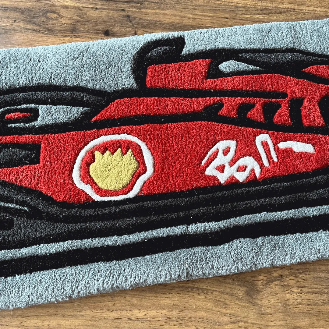 asphalt aura red race car rug detail Close-up detail of the Asphalt Aura rug featuring the nose of a red Formula 1 style race car with stylized white and yellow logos on a grey background.
