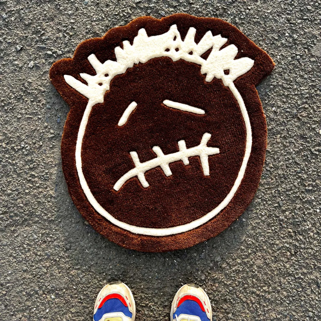 bruma mark rug lifestyle First-person POV lifestyle view of the Bruma Mark rug placed on a concrete street setting, showcasing the brown and white stylized face design alongside a pair of colorful sneakers at the bottom edge.