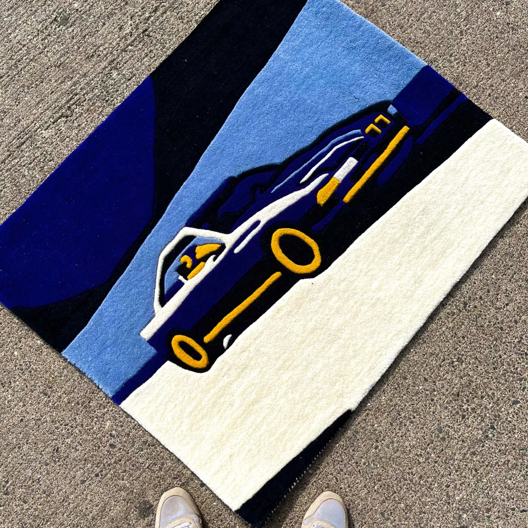 noctra veil rug lifestyle First-person angled lifestyle view of the Noctra Veil rug styled on a concrete street setting, showcasing the vibrant automotive design alongside a pair of sneakers at the bottom edge.