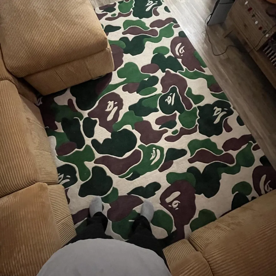 shadowfield camo terrain rug pov First-person point of view standing on the Shadowfield Camo Terrain rug, styled in a cozy living room next to a tan corduroy sectional sofa, emphasizing the scale of the bold camo pattern.