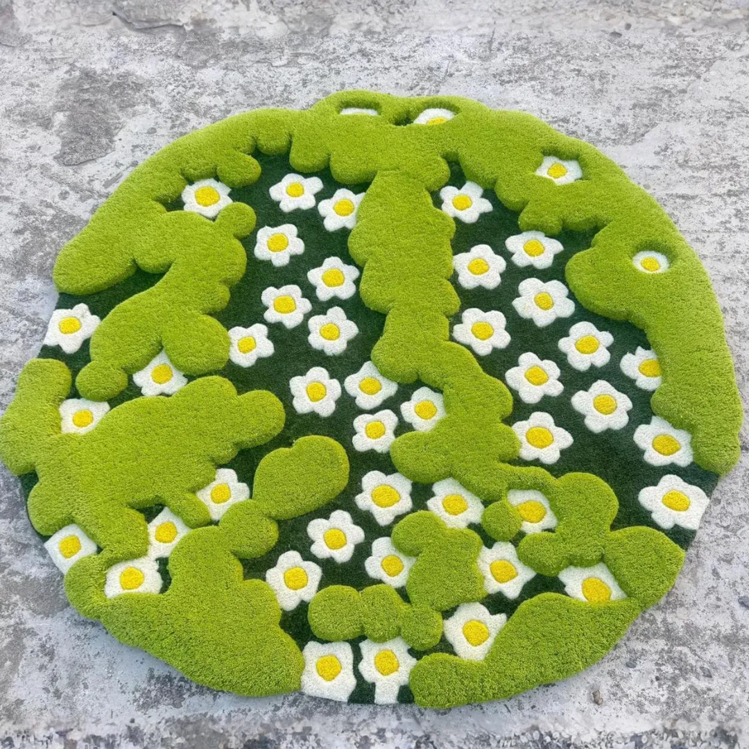 3d moss rug full overhead view Top-down view of the circular 3-D Moss Rug resting on a light grey concrete surface. The hand-tufted design features raised, bright green mossy shapes overlapping a dark green background filled with white and yellow daisy flowers.