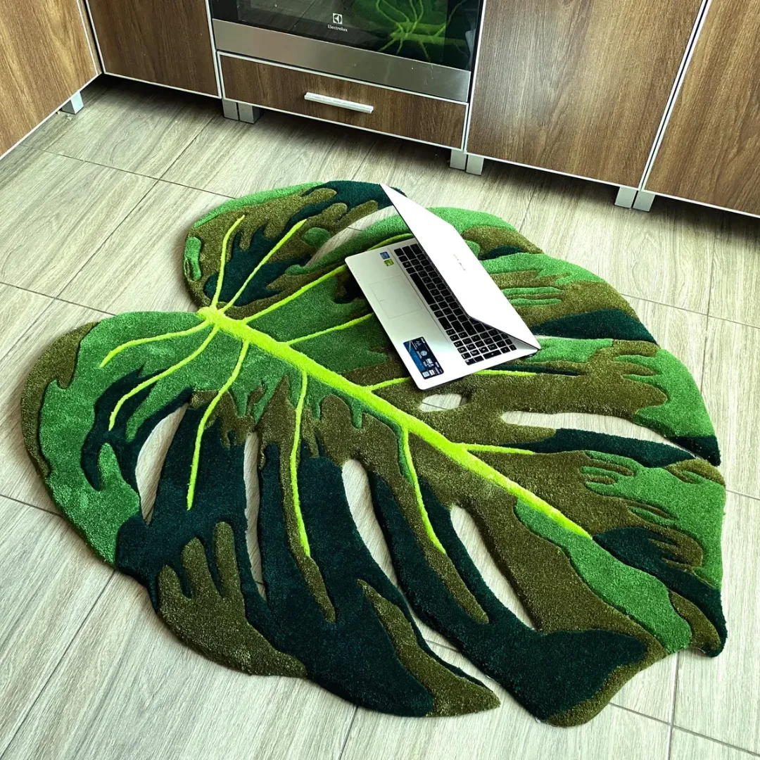monstera flux rug top view with laptop Top-down view of the tufted Monstera Flux Rug on a light tiled floor with a silver laptop resting on it for scale. The rug features a custom cut-out tropical leaf design in multiple shades of green.