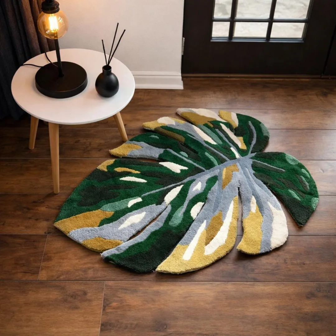 monstera tropical rug dark wood floor An angled view of the Monstera Tropical Rug displayed on a dark wood floor near a small wooden side table with a lit lamp. The sunlight highlights the thick, tufted texture and the intricate cut-out edges of the green, yellow, and grey leaf design.