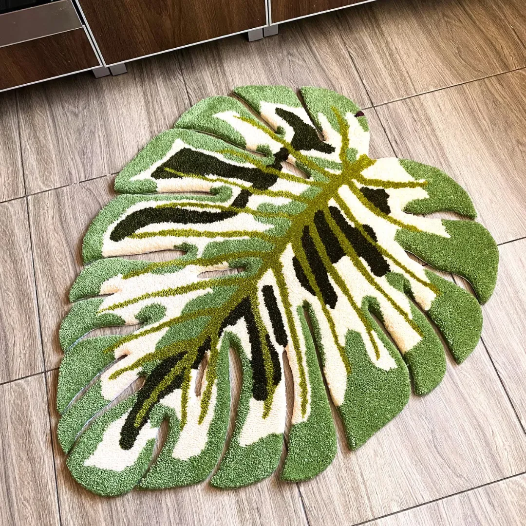 verdant monstera rug full view Full overhead view of the Verdant Monstera Rug laid flat on a wood-look tiled floor. The hand-tufted tropical leaf design features a white base with light green borders, a bright green central vein, and dark green shading.