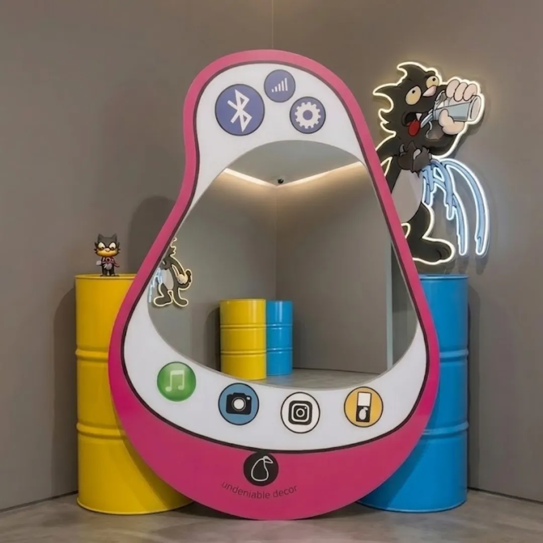 aura pear phone Interior styling view of the Aura Pear Phone Mirror displayed in a creative pop art studio space flanked by bright yellow and blue industrial barrels and glowing neon cartoon wall decor.