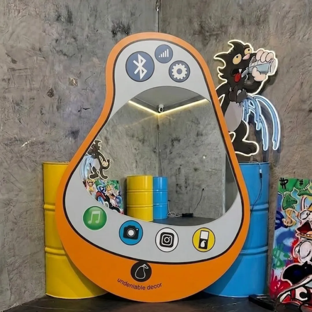 pear pop phone mirror pop art room styling Interior styling view of the Pear Pop Phone Mirror displayed in a creative pop art studio space flanked by bright yellow and blue industrial barrels and glowing neon cartoon wall decor.