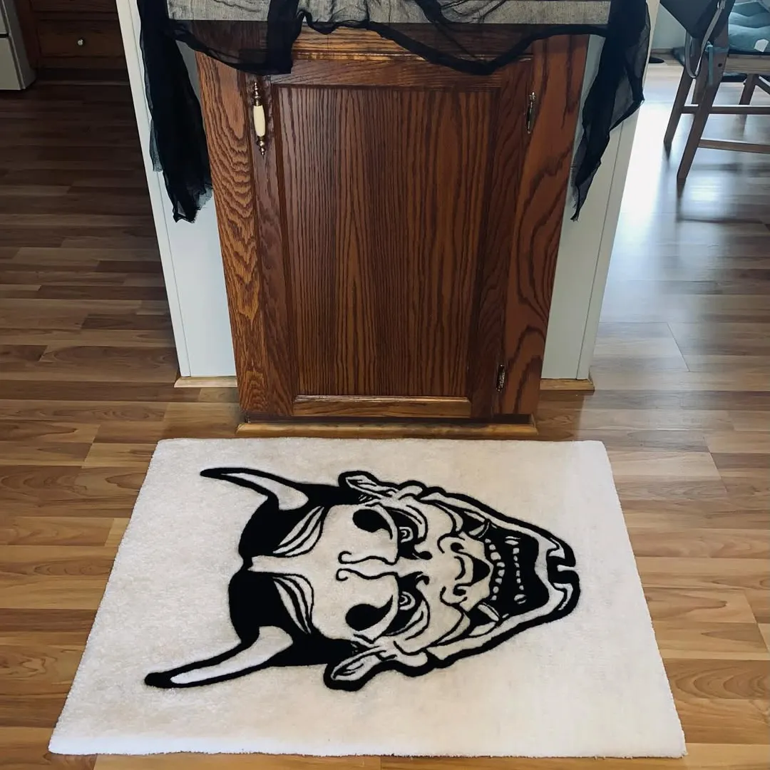 Rectangular white rug featuring a bold black Hannya mask design with sharp horns, fierce eyes, and a dramatic expression, placed on wooden flooring.