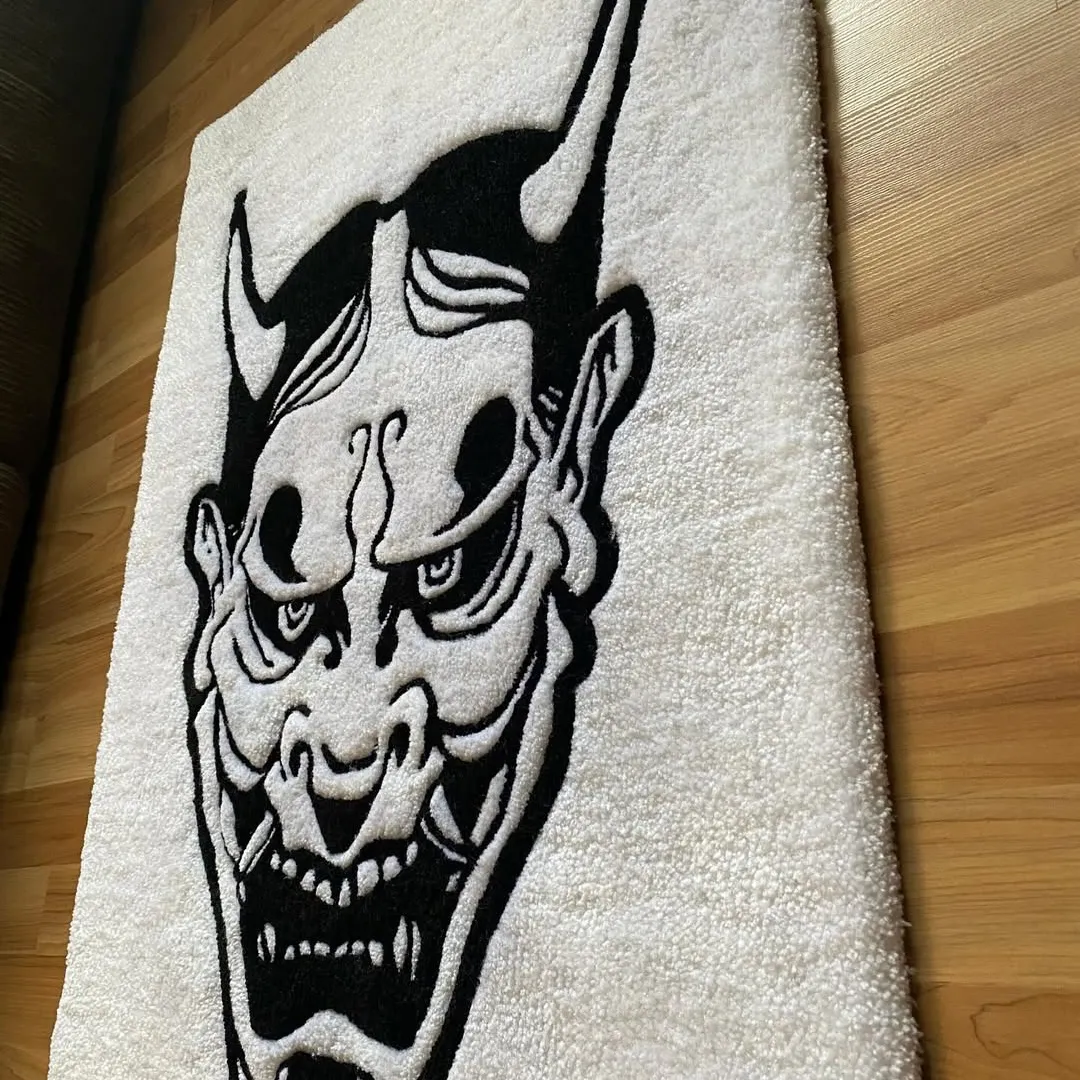Close-up shot of a white hand-tufted rug with a stylized black Hannya face pattern, emphasizing the texture and dramatic line work.