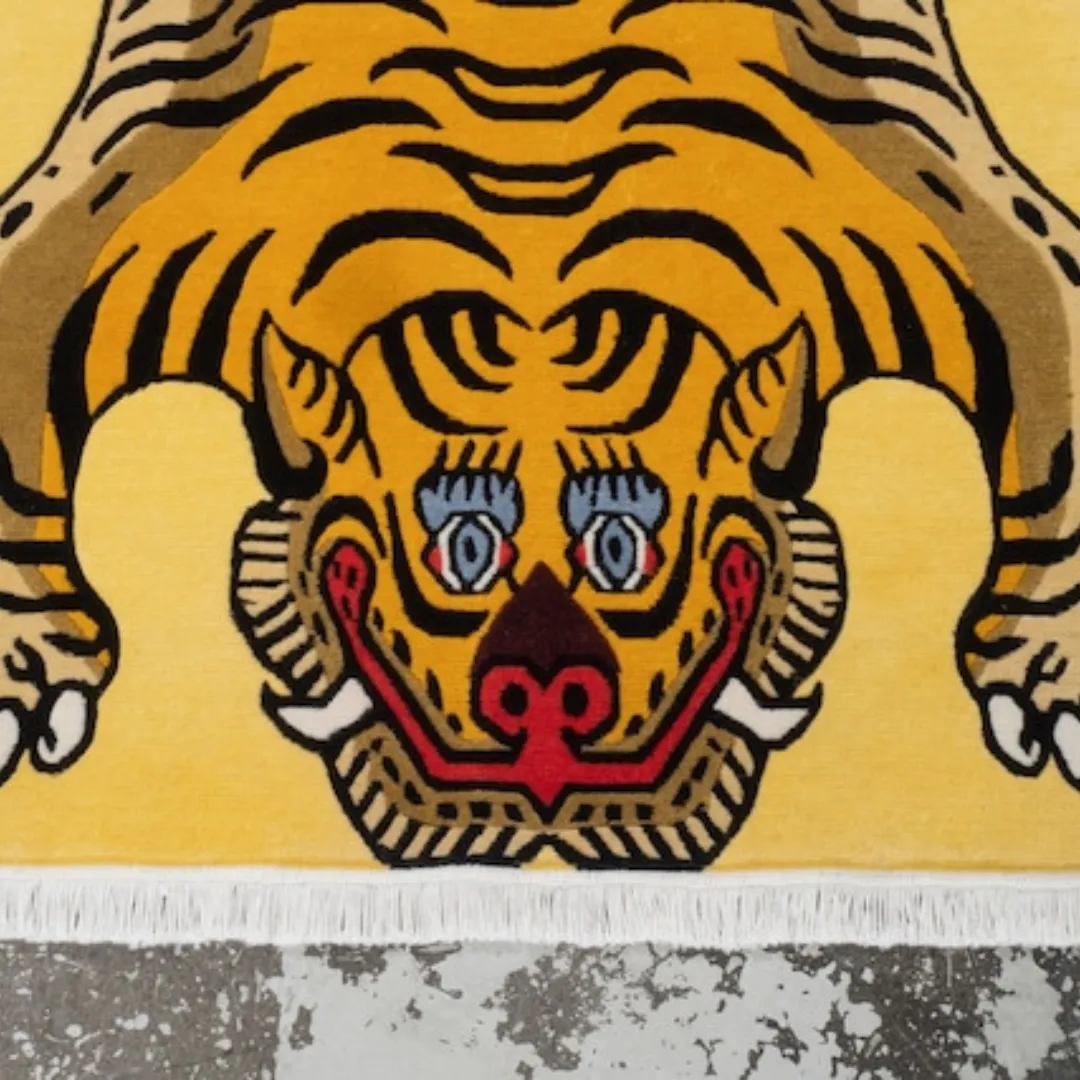 Hand tufted yellow Tibetan-style tiger rug with bold black stripes, red face details, and white fringe, displayed on a gray concrete floor.