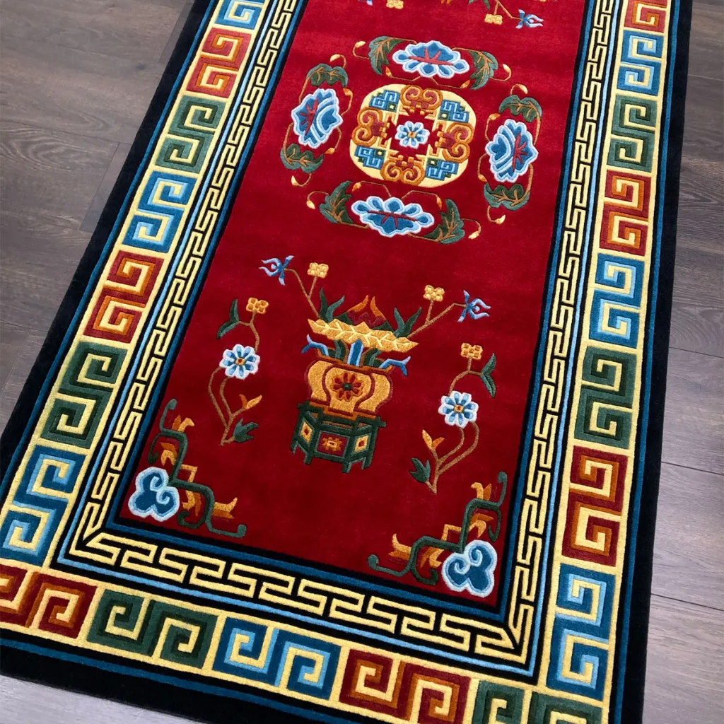 traditional buddhist floral design area rug Angled shot of the Lotus Throne area rug displaying traditional auspicious symbols and stylized floral art in bright colors against a rich maroon base.