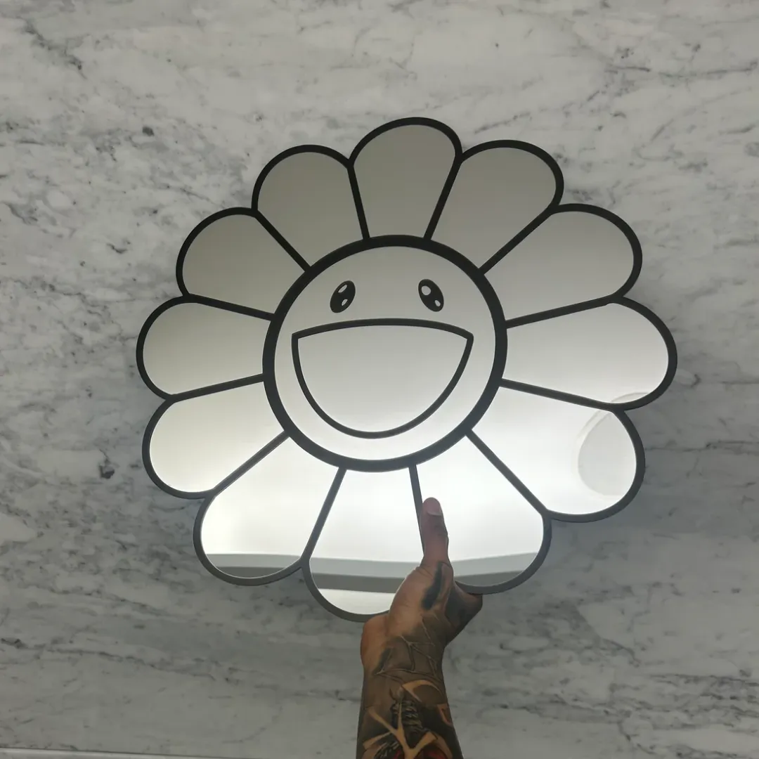 smiley daisy flower mirror hand held A hand holding up a daisy-shaped mirror with a bold black outlined smiley face in the center, set against a bright white marble background.