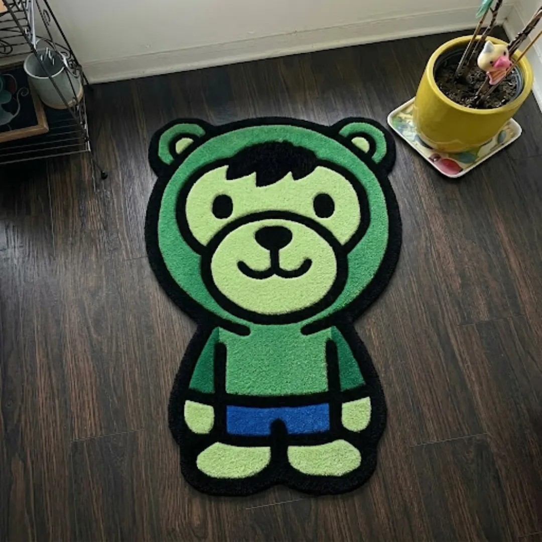 gamma fury rage shaped area rug product shot A top-down studio product photograph of a unique, character-shaped area rug featuring a green bear character wearing blue shorts and a hooded top, displayed on a dark wooden floor.
