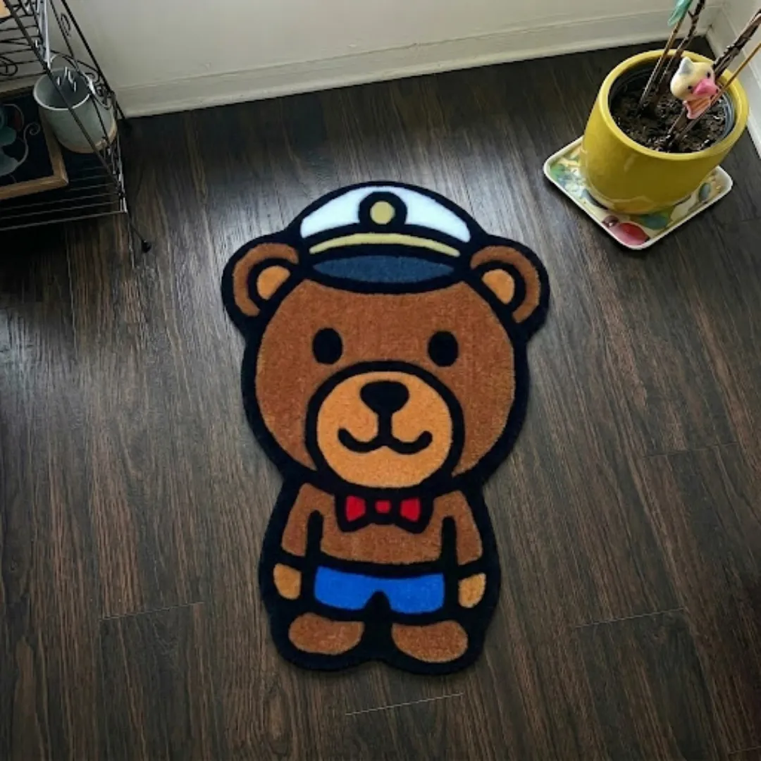 harbour line captain shaped area rug product shot A top-down studio product photograph of a character-shaped area rug featuring a cute brown bear wearing a captain's hat, red bowtie, and blue shorts, displayed on a wooden floor.