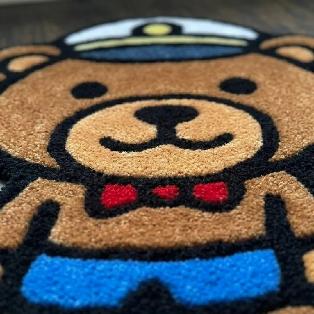 harbour line captain shaped area rug texture close up A macro close-up of the captain bear rug, focusing on the dense, tufted wool pile and the detailed graphic elements of the bear's face, bowtie, and blue shorts.