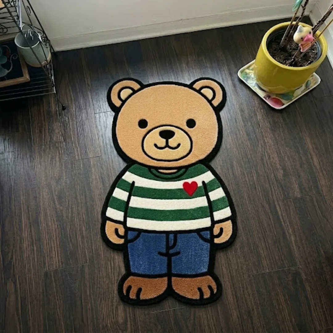 stormbound ape shaped area rug product shot A top-down studio product photograph of a unique, character-shaped area rug featuring a cute brown bear character in a green-and-white striped sweater with a red heart and blue jeans, displayed on a wooden floor.